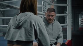 Male trainer sits on ring and explains fighting technique to female boxer. Woman listens coach and repeats exercises. Fighter prepares to match in gym. Physical activity and intensive workout. - Powered by Shutterstock - Get 15% off with code: PIKWIZARD15