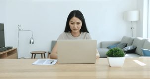Serious busy young Asian entrepreneur woman typing on laptop, sitting at large workplace table, working on business project at home, reading, watching content, touching chin, thinking