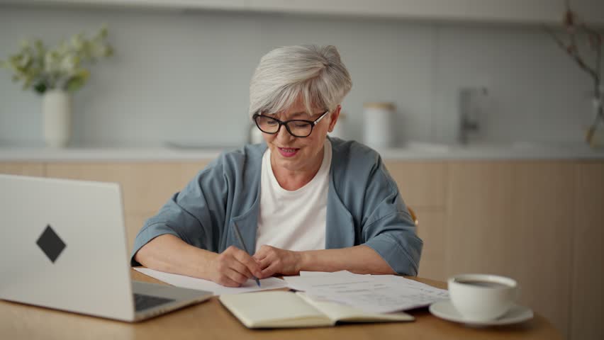Senior woman looking at laptop, watching online lesson, studying. Elderly grandma sitting at home at the kitchen is engaged in self-education. Self-education and learning for older people concept.