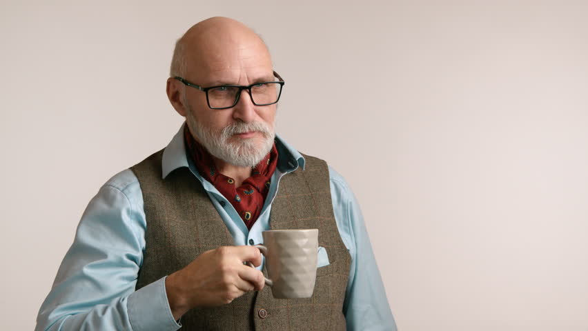 A contemplative senior man enjoying a cup of coffee while thoughtfully glancing at his watch, wearing a casual shirt and vest, against a soft beige backdrop. High quality 4k footage Royalty-Free Stock Footage #3425785223