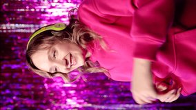 Young excited woman in pink hoodie listening music in yellow headphones and dancing around violet background. Vertical video.