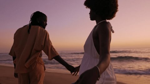 Silhouetted image of a young African couple in stylish attire walking along the beach during a beautiful sunset, displaying affection and togetherness.の動画素材
