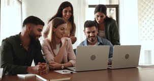 Happy multiracial mates laugh while collaborating on new project in office with laptops. Group of diverse employees using notebooks, have fun, watch online content, work together, engaged in teamwork