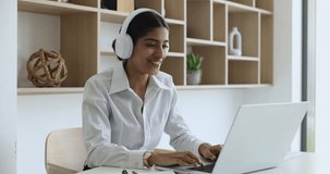 Positive beautiful young Indian manager woman in headphones talking on online video conference call, speaking at laptop screen, smiling, enjoying internet business communication