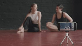 Rack focused shot of two young Caucasian female dancers sitting on parquet floor and talking while recording choreography video class on smartphone standing on tripod in foreground