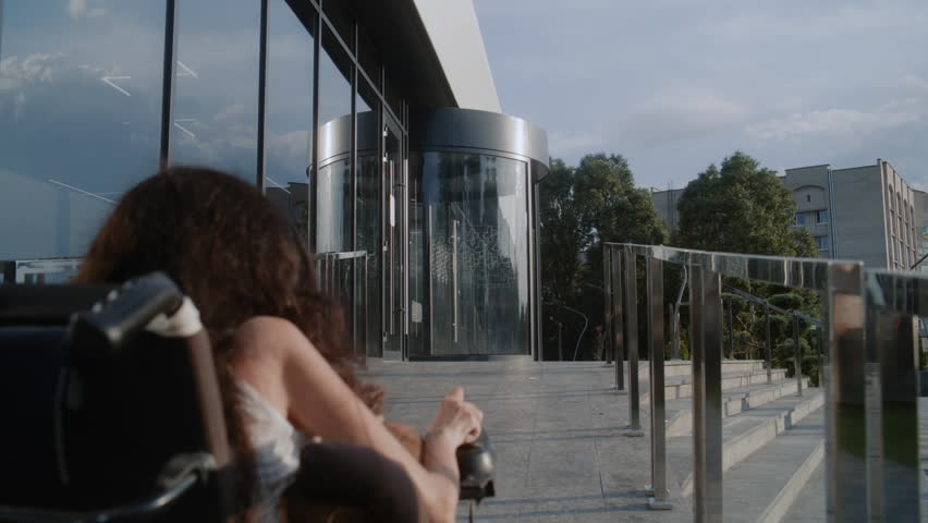 Woman with physical disability rides into clinic entrance on motorized wheelchair. Male doctor meets female patient, helps her enter through revolving doors. Hospital or modern medical center outside.