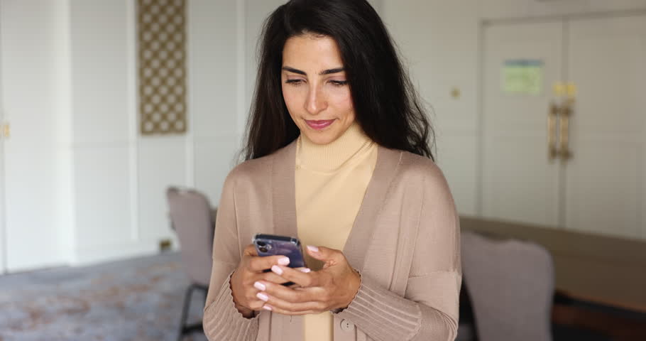 Attractive Latina woman holding mobile phone, read message, get good news, smile, enjoy spending time in social network. Positive female enjoy e-dating services at home using new app on smart phone Royalty-Free Stock Footage #3426440521