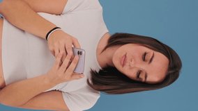 Vertical video, Beautiful young brunette woman dressed in white top hold using mobile cell phone isolated on blue background in studio