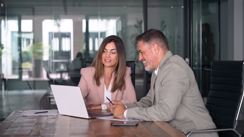 Smiling middle aged professional business woman executive working looking at laptop computer discussing digital data with male colleague at office meeting planning corporate strategy.