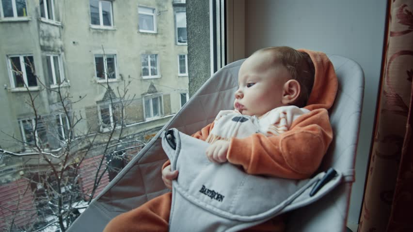 A pensive baby gazes out a window while snugly seated in a modern bouncer, enveloped by the warmth of a hooded garment. The outside world presents a stark contrast with its cool, urban landscape. Royalty-Free Stock Footage #3426618689