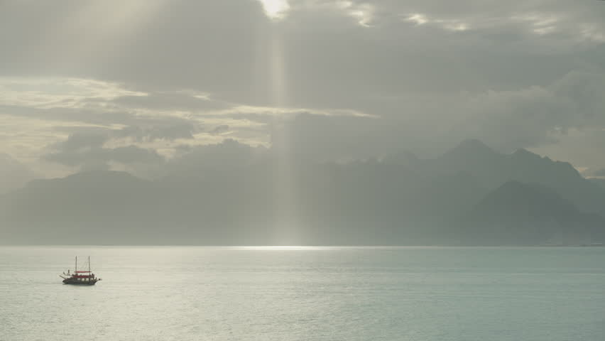 A boat in the distance on the sea, and a sunbeam through the clouds, resembling a spotlight, illuminates the path on the water. Mountains in the distance. Time-lapse. Royalty-Free Stock Footage #3426724293