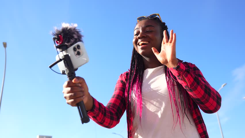 African woman waving while vlogging using mobile phone and microphone outdoors