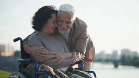 Husband walking with wife in wheelchair. Senior family couple in city park near pond. Elderly man hugging support woman showing love care, they talk smiling. Living with physical disability concept. - Powered by Shutterstock - Get 15% off with code: PIKWIZARD15