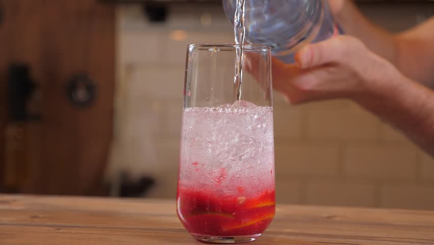 Close-up of the bartender pouring soda or water into a glass with berry syrup and ice, the process of making berry lemonade in the bar.