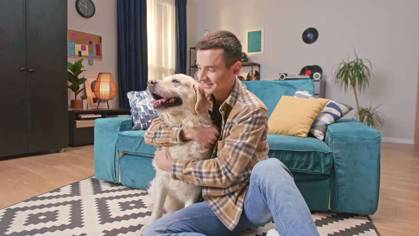 Zooming in on attractive Caucasian man with checkered shirt sitting on carpet and hugging his dog. Young owner petting labrador retriever and cheerfully smiling on camera. Animal concept.