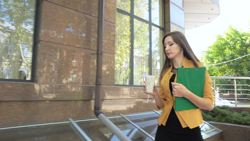 Young woman walking down street with green folder and drinking coffee