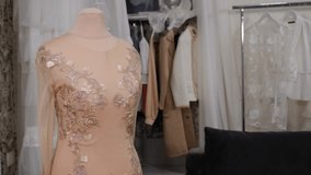 Close-up of a beautiful beige openwork dress on a mannequin in the atelier, in the background there are hangers with clothes.