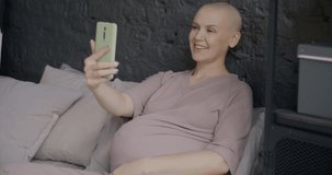 Future mother making online video call talking laughing waving hand using smartphone lying in bed at home. Communication and pregnancy concept.