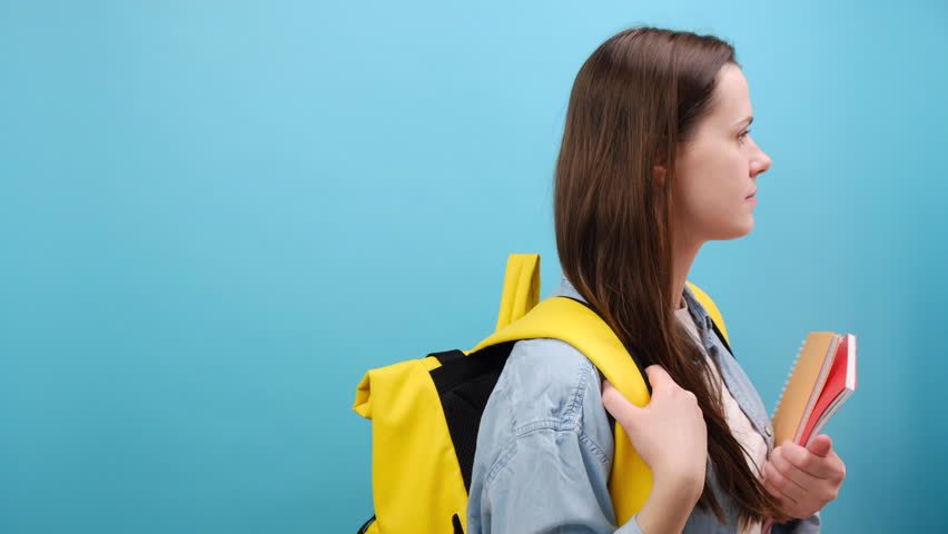 Side view of cheerful cute young woman student in shirt with backpack looking aside, posing isolated over blue studio background with copy space. Education in high school university college concept Royalty-Free Stock Footage #3427053673