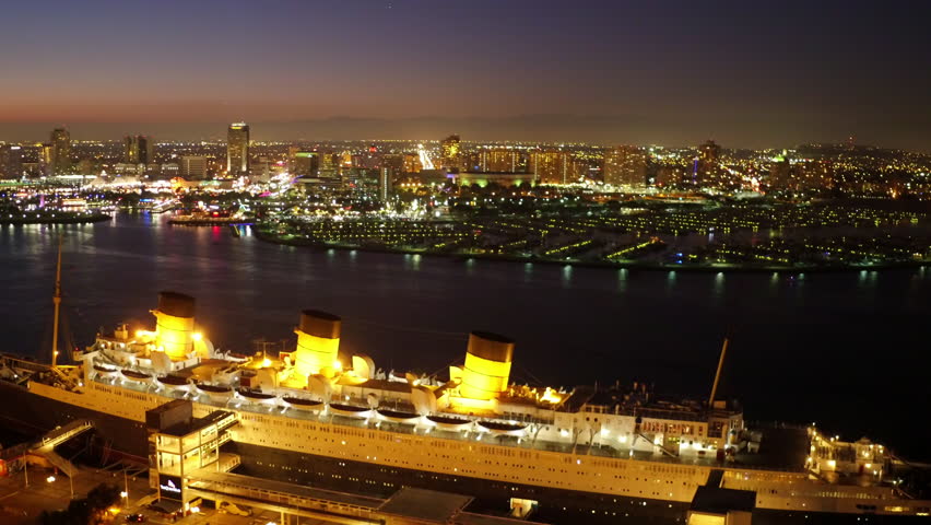 Aerial Panning Shot Of The Queen Mary Cruise Ship Moored In Sea By Illuminated City Against Sky At Night - Long Beach, California