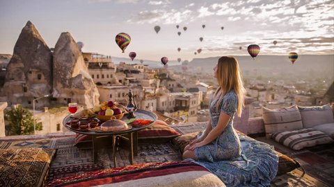 Woman sits on the top of roof and enjoys the morning view on Goreme park with air balloons fly by