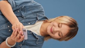 Vertical video, Laughing young woman dressed in denim shirt having fun looking at mobile phone isolated on blue backgroundin studio