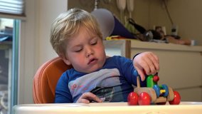 Two-year-old cute blond boy sits on a highchair with a table in the room and playing with colorful toys
