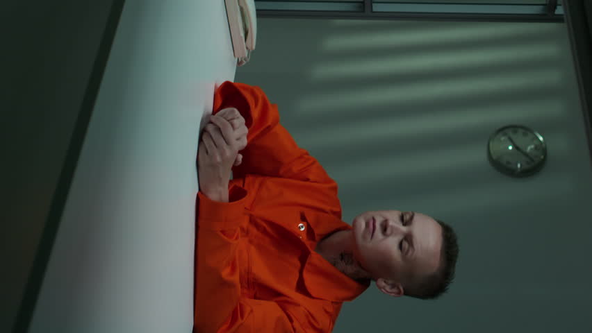 Vertical shot of arrested short-haired woman in orange prison uniform sitting at table in interrogation room and waiting for interview