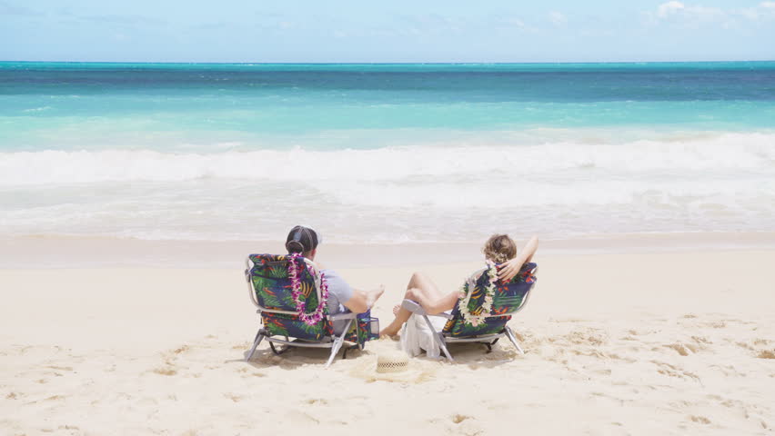 Luxury tropical vacation resort lifestyle background. Two tourists relaxing in beach chairs. Back view of man and woman enjoying blue ocean. Travelers sun bathing at beach on summer holiday Hawaii USA