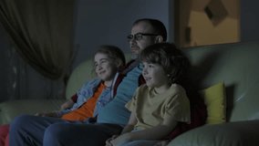 Happy Smiling Family With Children Sitting On Sofa In Living Room Evening Watching TV Together. Father With Son and Daughter Watching Movie at Home in Living Room at Night. Togetherness Parenthood.