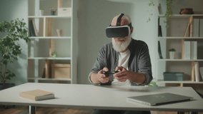 An Old Man in VR Glasses Plays Computer Games With a Joystick, Sitting at the table.A Modern Elderly Man Plays PlayStation. Augmented Reality and Gaming for Seniors.