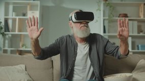 A Modern Old Man in VR Glasses Examines and Touches Everything Around. An Elderly Man in VR Glasses Sitting in the Sofa with a Popcorn.Virtual Reality Games for the Elderly.