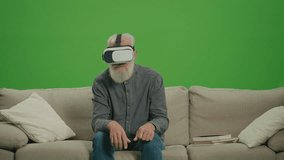Green Screen. An Old Man With Gray Beard in VR Glasses Examines and Touches Everything Around, Sitting on a Sofa. Medical Applications of VR for the Elderly.