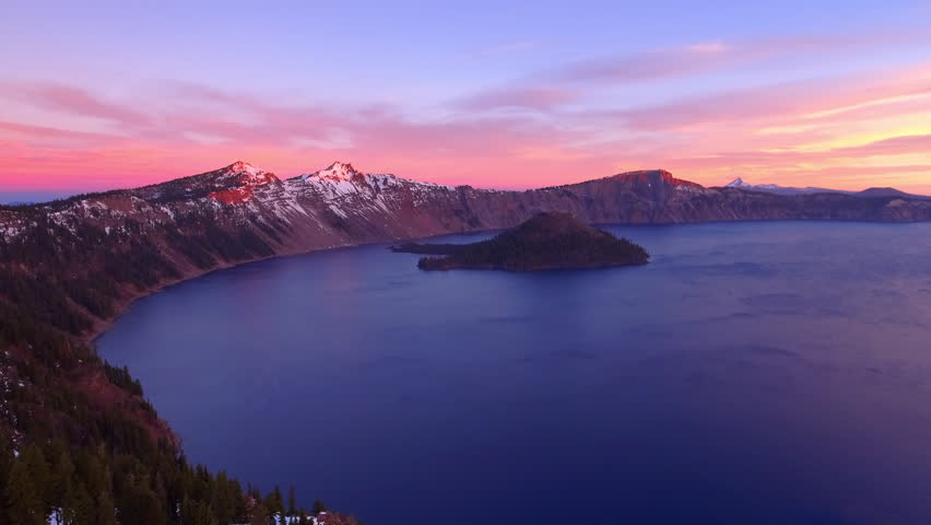 Aerial Scenic View Of Crater Lake National Park, Drone Flying Backwards Over Trees On Snowy Mountains During Sunset