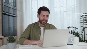 Caucasian bearded man in casual outfit using modern laptop and internet connection for having online interview. Handsome guy discussing work details with hiring company at domestic workplace.
