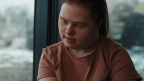 Pensive down syndrome woman sitting next to window and looking away