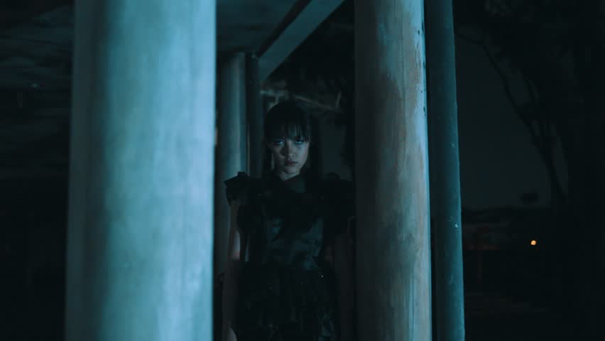 A young woman in a gothic black dress and pigtails stands between pillars in a dark, blue-tinted environment at night. She stares intently at the camera with a creepy, unsettling expression.