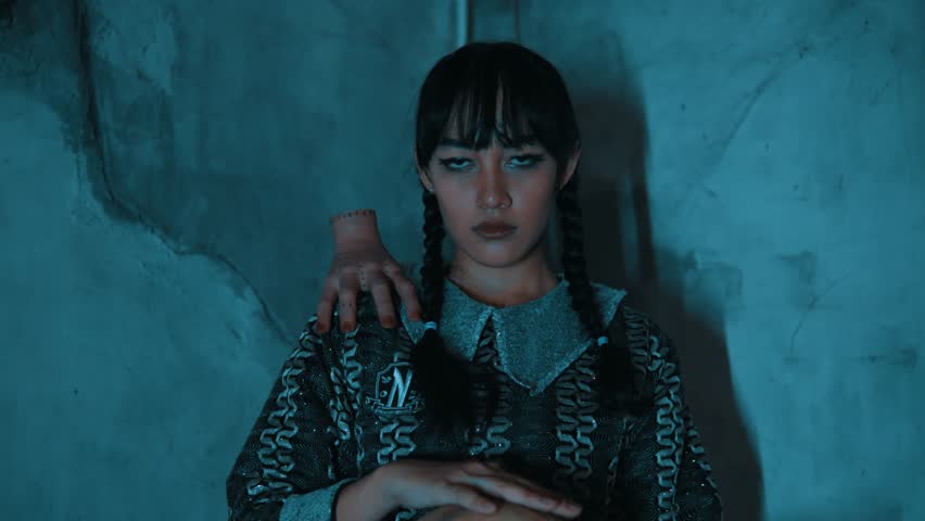 A zoom-out shot starting with a close-up of a creepy gothic girl staring at the camera. A severed hand is on her shoulder, and she holds a skull. The shot reveals her in a dark, derelict room.