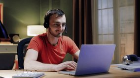 Male student wear headset e-learning studying with online tutor teacher talk conference calling do video chat learn language make notes sitting at home office desk look at laptop computer