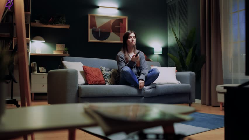 Attractive interested woman enjoying favorite TV program while relaxing on couch. Sitting in front of modern large-screen high-definition TV, contented young female watching feel-good drama set. Royalty-Free Stock Footage #3427863487