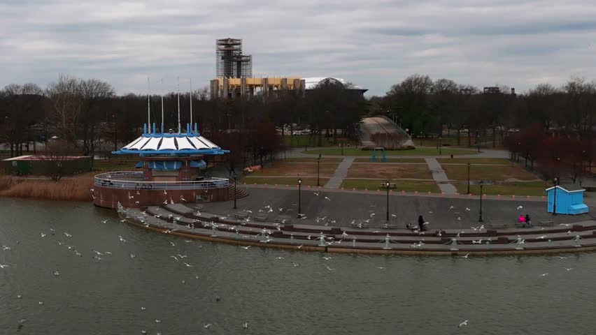 An aerial view of Meadow Lake in Flushing Meadows Corona Park in NY on a cloudy day. The camera is stationary as seagulls fly in slow motion and structures from the worlds fair are in the background.