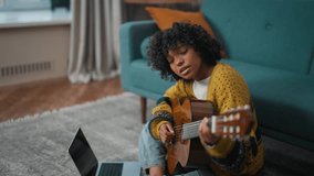 Young girl learning to play guitar. African american teenager girl study to playing the guitar sitting on floor at home with a laptop. Online-education, singing, create hobby practice concept.
