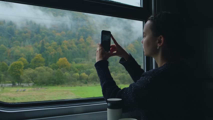 Young woman in train. Girl takes photo of mountain landscape outside the window while sitting inside transport. Passenger or traveler takes photos with phone. Woman taking pictures of Carpathian mount