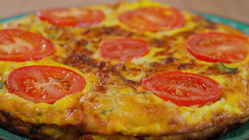 Healthy vegetarian food. Ready omelette with tomatoes, frittata rotating on a round plate, wooden table. Close-up, side view.