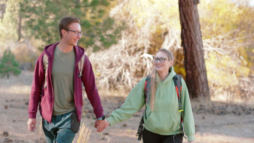 Happy couple enjoying a nature hike, holding hands and smiling in a sunlit forest, showcasing a shared love for outdoor activities. Slow Motion, Camera 4K RAW.  Royalty-Free Stock Footage #3428199211