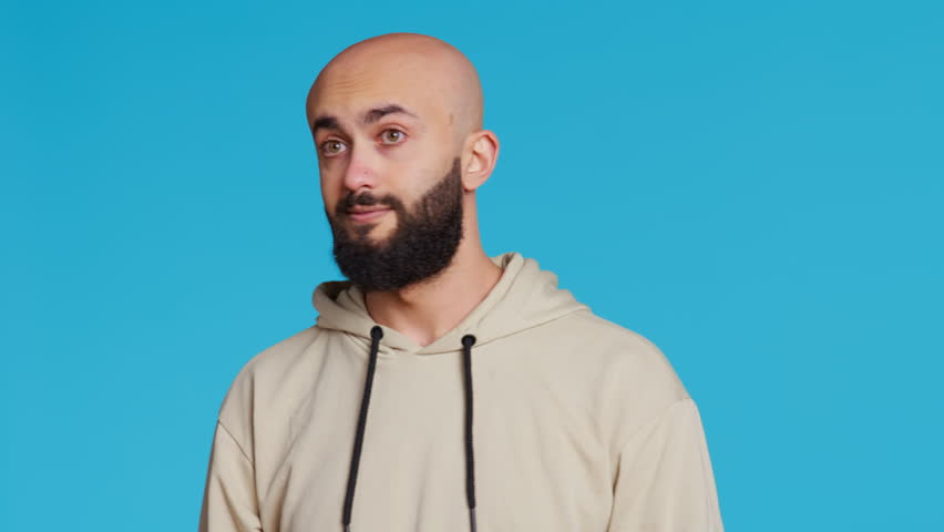 Middle eastern guy greeting people in front of camera, waving hello and smiling over blue background. Young person posing with confidence in studio, feeling confident and joyful. Camera 2.