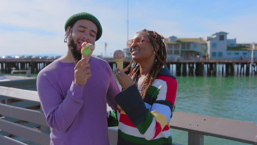 A black man in a green beanie and black woman with braided hair laugh while eating ice cream on a sunny pier overlooking the sea. Slow Motion, Camera 4K RAW.  Royalty-Free Stock Footage #3428218527