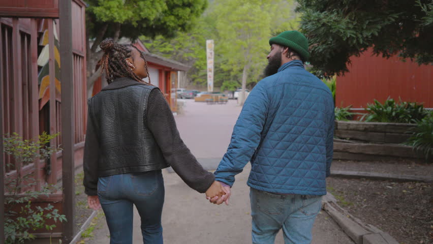 Back view of a diverse African American couple holding hands and walking down a peaceful street, with rustic cabins and lush greenery around. Slow Motion, Camera 4K RAW. 