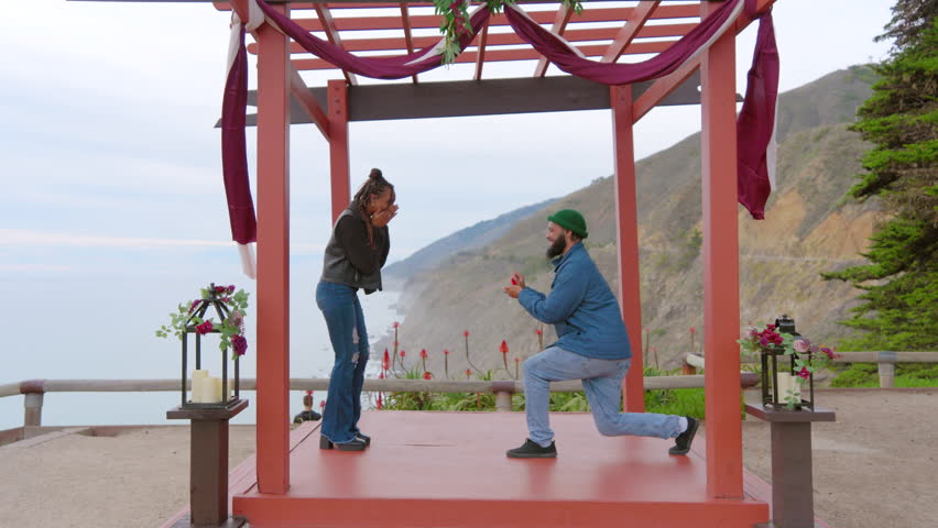 African American man kneels in a surprise proposal to his partner at a scenic overlook, with a stunning cliffside ocean view setting the scene. Slow Motion, Camera 4K RAW. 