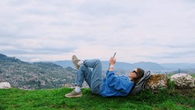 A young happy smiling girl lying on the green grass on the top of hill overlooking a European city. Relaxed woman traveler using smartphone, making photo and video of her journey.